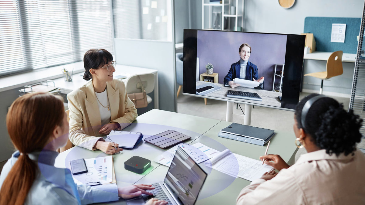 Three colleagues around a meeting-room table with a Coolpo device between them, a remote teammate mid-sentence on the wall screen