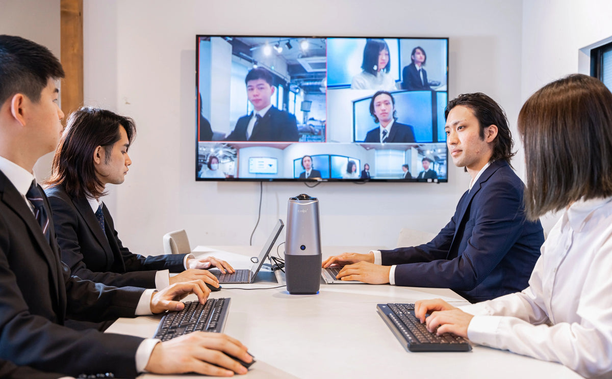 Four colleagues in a meeting room with a Coolpo AI Huddle PANA on the table, the wall screen showing remote participants and the camera's 360-degree panoramic strip