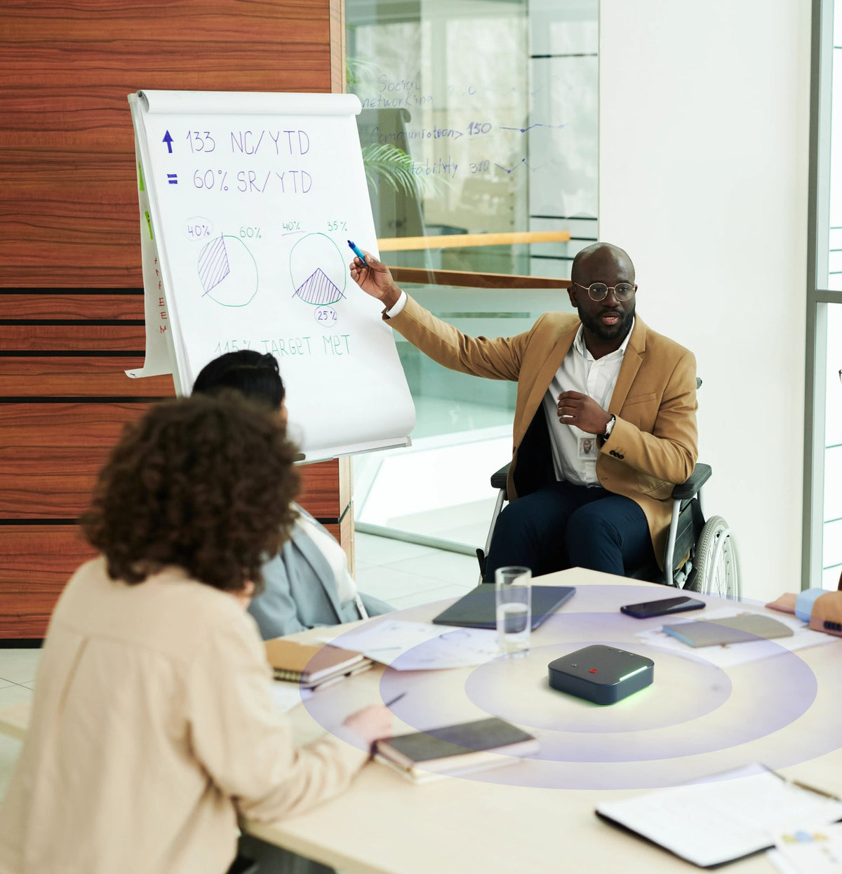 A team meeting in progress: one colleague presenting figures at a flip chart while others follow from around the table, a Coolpo device between them